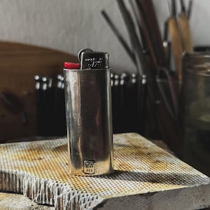 May include: A silver lighter with a red top and the text "made in USA 9172" on the side. The lighter is sitting on a textured, gray surface. The lighter is stamped with "WM 925".