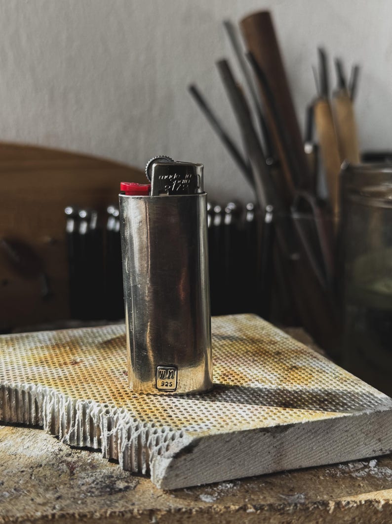 May include: A silver lighter with a red top and the text "made in USA 9172" on the side. The lighter is sitting on a textured, gray surface. The lighter is stamped with "WM 925".