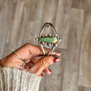 May include: A silver-toned cuff bracelet with a large, oval turquoise stone at the center. The turquoise is a vibrant green with brown matrix. The bracelet has decorative silver accents and is held by a hand.