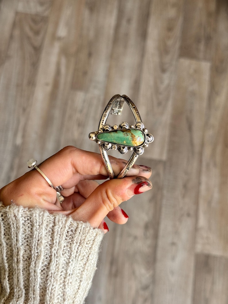 May include: A silver-toned cuff bracelet with a large, oval turquoise stone at the center. The turquoise is a vibrant green with brown matrix. The bracelet has decorative silver accents and is held by a hand.