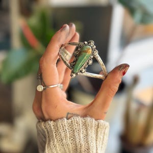 May include: Silver cuff bracelet featuring a large, teardrop-shaped turquoise stone. The stone is set in a decorative bezel with intricate silverwork. The bracelet has a split design and is held in a hand.