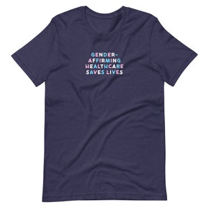 May include: Navy blue t-shirt with the text "GENDER-AFFIRMING HEALTHCARE SAVES LIVES" in pastel colors. The text is stacked vertically. The shirt is a short-sleeved crew neck.