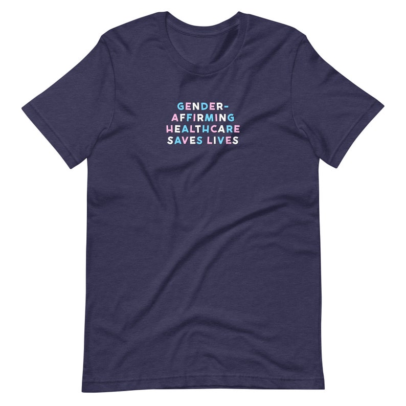 May include: Navy blue t-shirt with the text "GENDER-AFFIRMING HEALTHCARE SAVES LIVES" in pastel colors. The text is stacked vertically. The shirt is a short-sleeved crew neck.
