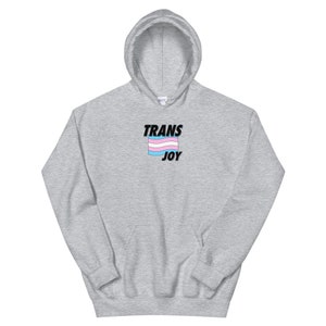 以下が含まれることがあります： 前面にトランスジェンダーのプライドフラッグと「TRANS JOY」の文字がプリントされたグレーのパーカー