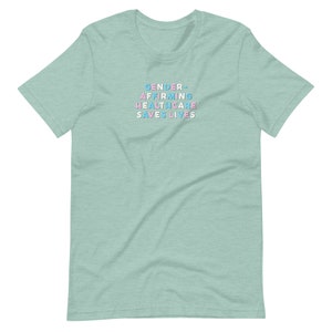 May include: A light green t-shirt with the text "GENDER-AFFIRMING HEALTHCARE SAVES LIVES" in pastel blue, pink, and white block letters. The shirt is a crew neck style and has short sleeves.