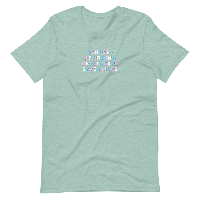 May include: A light green t-shirt with the text "GENDER-AFFIRMING HEALTHCARE SAVES LIVES" in pastel blue, pink, and white block letters. The shirt is a crew neck style and has short sleeves.