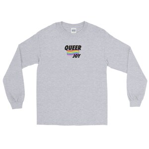 以下が含まれることがあります： レインボーフラッグのグラフィックと黒い文字で「QUEER JOY」と書かれたヘザーグレーのロングスリーブTシャツです。