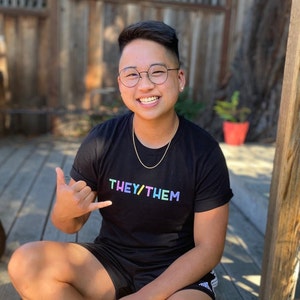 May include: A person wearing a black t-shirt with the text "THEY/THEM" in rainbow colours. The person is smiling and has short black hair. They are wearing round glasses and a gold chain necklace.