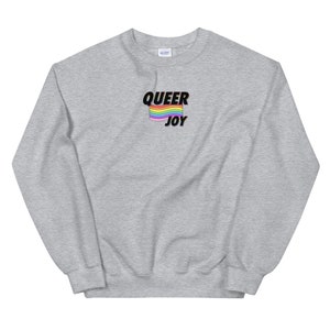以下が含まれることがあります： 「QUEER JOY」の文字と虹色の旗のデザインがプリントされた、ライトグレーのスウェットシャツ。クルーネックのスタイルで、柔らかい素材で作られているようです。