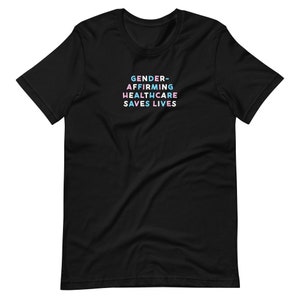 May include: Black t-shirt with the text "GENDER-AFFIRMING HEALTHCARE SAVES LIVES" in a stacked format. The text is in a pastel color scheme of blue, pink, and white. The shirt has short sleeves and a crew neck.