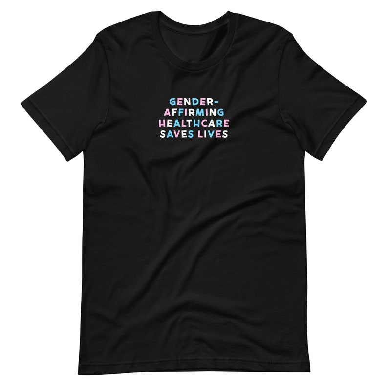 May include: Black t-shirt with the text "GENDER-AFFIRMING HEALTHCARE SAVES LIVES" in a stacked format. The text is in a pastel color scheme of blue, pink, and white. The shirt has short sleeves and a crew neck.