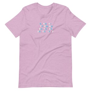 May include: A light pink t-shirt with the text "GENDER AFFIRMING HEALTHCARE SAVES LIVES" in a stacked, colorful font. The text is in shades of blue, pink, and white. The shirt has short sleeves and a crew neck.
