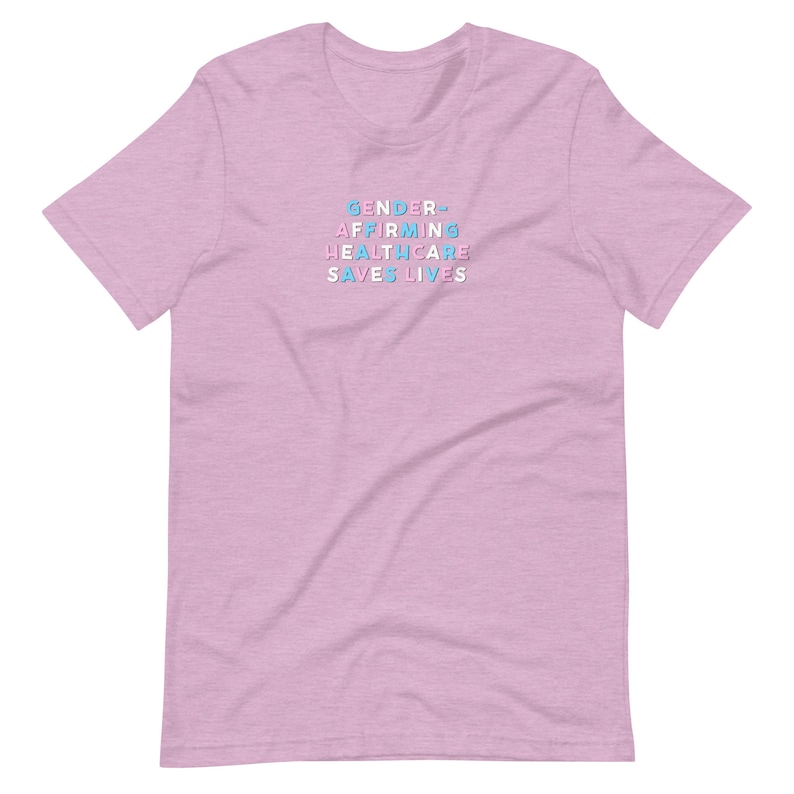 May include: A light pink t-shirt with the text "GENDER AFFIRMING HEALTHCARE SAVES LIVES" in a stacked, colorful font. The text is in shades of blue, pink, and white. The shirt has short sleeves and a crew neck.