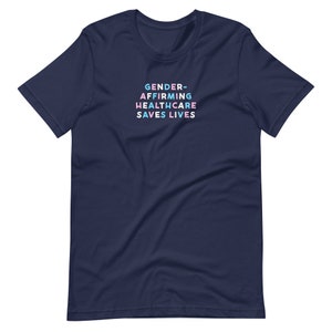 May include: Navy blue t-shirt with the text "GENDER-AFFIRMING HEALTHCARE SAVES LIVES" in a stacked, colorful font. The text is in shades of blue, pink, and white. The shirt is a short-sleeved crew neck.
