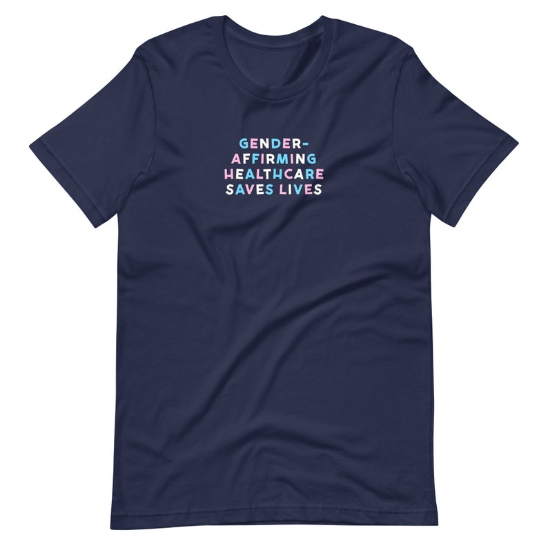 May include: Navy blue t-shirt with the text "GENDER-AFFIRMING HEALTHCARE SAVES LIVES" in a stacked, colorful font. The text is in shades of blue, pink, and white. The shirt is a short-sleeved crew neck.