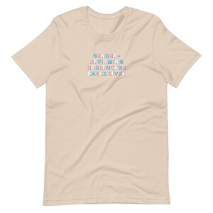 May include: A beige t-shirt with the text "GENDER-AFFIRMING HEALTHCARE SAVES LIVES" in blue, pink, and white block letters. The shirt has short sleeves and a crew neck.
