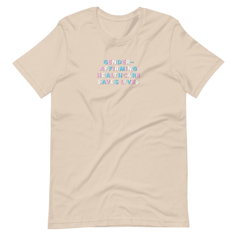 May include: A beige t-shirt with the text "GENDER-AFFIRMING HEALTHCARE SAVES LIVES" in blue, pink, and white block letters. The shirt has short sleeves and a crew neck.
