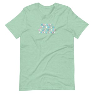 May include: A light green t-shirt with the text "GENDER-AFFIRMING HEALTHCARE SAVES LIVES" in pastel blue, pink, and white lettering. The shirt is a short-sleeved crew neck style.