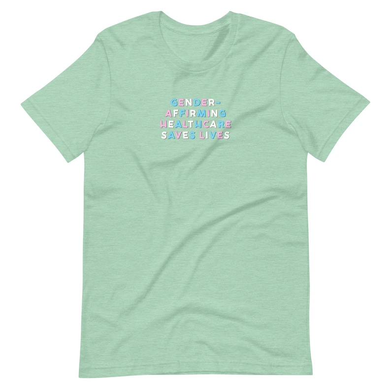 May include: A light green t-shirt with the text "GENDER-AFFIRMING HEALTHCARE SAVES LIVES" in pastel blue, pink, and white lettering. The shirt is a short-sleeved crew neck style.