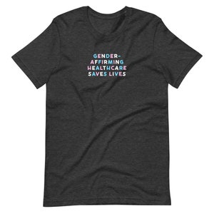 May include: Dark gray t-shirt with the text "GENDER-AFFIRMING HEALTHCARE SAVES LIVES" in a colorful, stacked font. The text is in shades of blue, pink, and white. The shirt is made of a soft material.