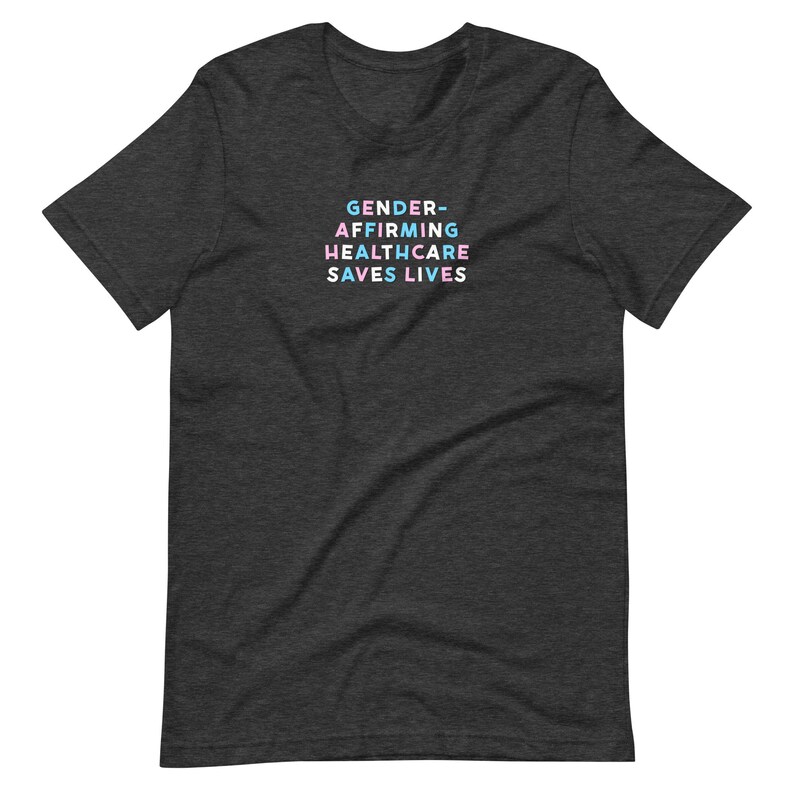 May include: Dark gray t-shirt with the text "GENDER-AFFIRMING HEALTHCARE SAVES LIVES" in a colorful, stacked font. The text is in shades of blue, pink, and white. The shirt is made of a soft material.