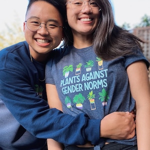 Könnte beinhalten: Zwei Personen lächeln und umarmen sich. Die Person im Vordergrund trägt ein dunkelblaues T-Shirt mit dem Text "Plants Against Gender Norms" und einer Grafik von Topfpflanzen.