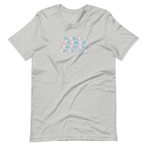 May include: A light gray t-shirt with the text "GENDER-AFFIRMING HEALTHCARE SAVES LIVES" in blue, pink, and light blue. The shirt is a short-sleeved crew neck.