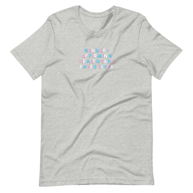 May include: A light gray t-shirt with the text "GENDER-AFFIRMING HEALTHCARE SAVES LIVES" in blue, pink, and light blue. The shirt is a short-sleeved crew neck.