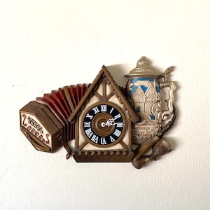 May include: A decorative wall hanging featuring a cuckoo clock, an accordion, and a beer stein. The clock has a black face with white Roman numerals. The accordion is brown and red. The beer stein is blue and white with a pipe.