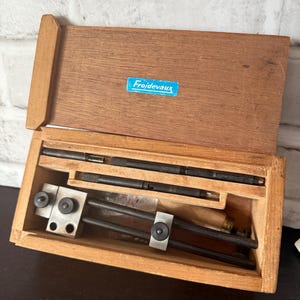 May include: A wooden storage box with a hinged lid featuring a blue label with the text Froidevaux. Inside, the box contains a set of metal precision tools, including two thin black rods and a sliding caliper-style instrument with black knobs.