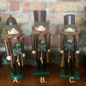 May include: Three wooden nutcracker figurines with brown hats and coats. Each nutcracker has a different design on their chest, including a clock, a bird, and a birdhouse. The nutcrackers are standing on green bases labeled A, B, and C.
