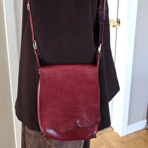 Puede incluir: Bolso bandolera de cuero burdeos con cierre de solapa y correa ajustable. El bolso tiene forma rectangular con bordes redondeados y un pequeño logotipo en la parte delantera. La correa está sujeta con anillos metálicos plateados.