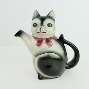 Könnte beinhalten: Eine Keramik-Teekanne in Form einer Katze, mit einem schwarz-weißen Körper, grünen Augen und einer rosa Schleife. Die Teekanne hat einen schwarzen Griff und Ausguss und ist als sitzende Katze gestaltet.