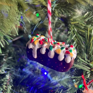 Peut inclure: Une d&eacute;coration de No&euml;l en forme de g&acirc;teau, de couleur marron et blanche, avec des vermicelles color&eacute;s et un gla&ccedil;age blanc.