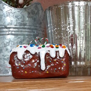 Peut inclure: Une d&eacute;coration de g&acirc;teau en forme de g&acirc;teau brun et blanc avec un gla&ccedil;age blanc et des p&eacute;pites color&eacute;es. La d&eacute;coration est orn&eacute;e d'un ruban rouge et blanc.