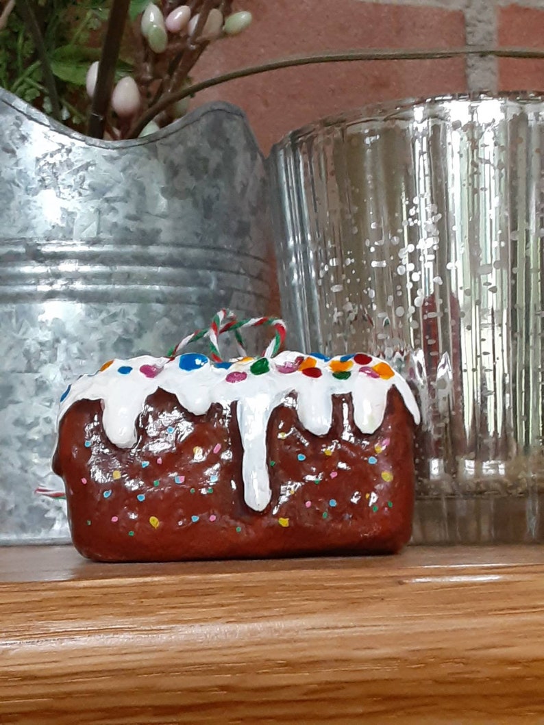 Peut inclure: Une d&eacute;coration de g&acirc;teau en forme de g&acirc;teau brun et blanc avec un gla&ccedil;age blanc et des p&eacute;pites color&eacute;es. La d&eacute;coration est orn&eacute;e d'un ruban rouge et blanc.