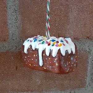 Peut inclure: Une d&eacute;coration de g&acirc;teau en forme de g&acirc;teau brun et blanc avec des p&eacute;pites color&eacute;es et un gla&ccedil;age blanc. La d&eacute;coration est suspendue &agrave; une ficelle rouge et verte.