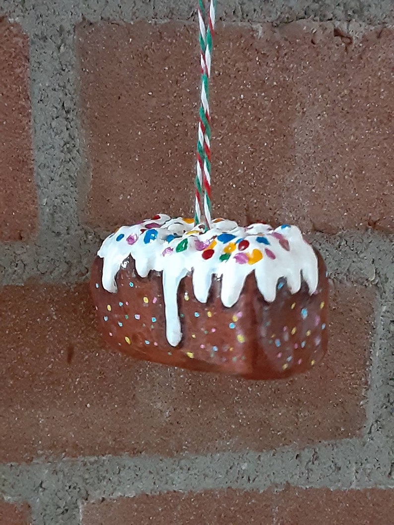 Peut inclure: Une d&eacute;coration de g&acirc;teau en forme de g&acirc;teau brun et blanc avec des p&eacute;pites color&eacute;es et un gla&ccedil;age blanc. La d&eacute;coration est suspendue &agrave; une ficelle rouge et verte.