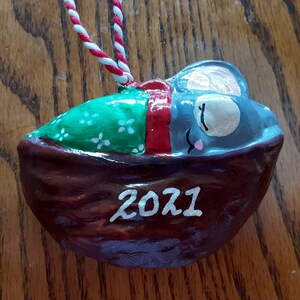 May include: A hand-painted ceramic ornament shaped like a walnut shell with a sleeping mouse inside. The mouse is gray with a pink nose and closed eyes. The shell is painted brown with white dots and the year 2021 is written on the front.
