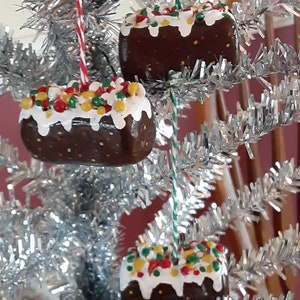 Peut inclure: Trois d&eacute;corations de No&euml;l en forme de g&acirc;teaux au chocolat avec un gla&ccedil;age blanc et des vermicelles color&eacute;s. Les d&eacute;corations sont suspendues &agrave; un sapin de No&euml;l en tinsel argent&eacute;.
