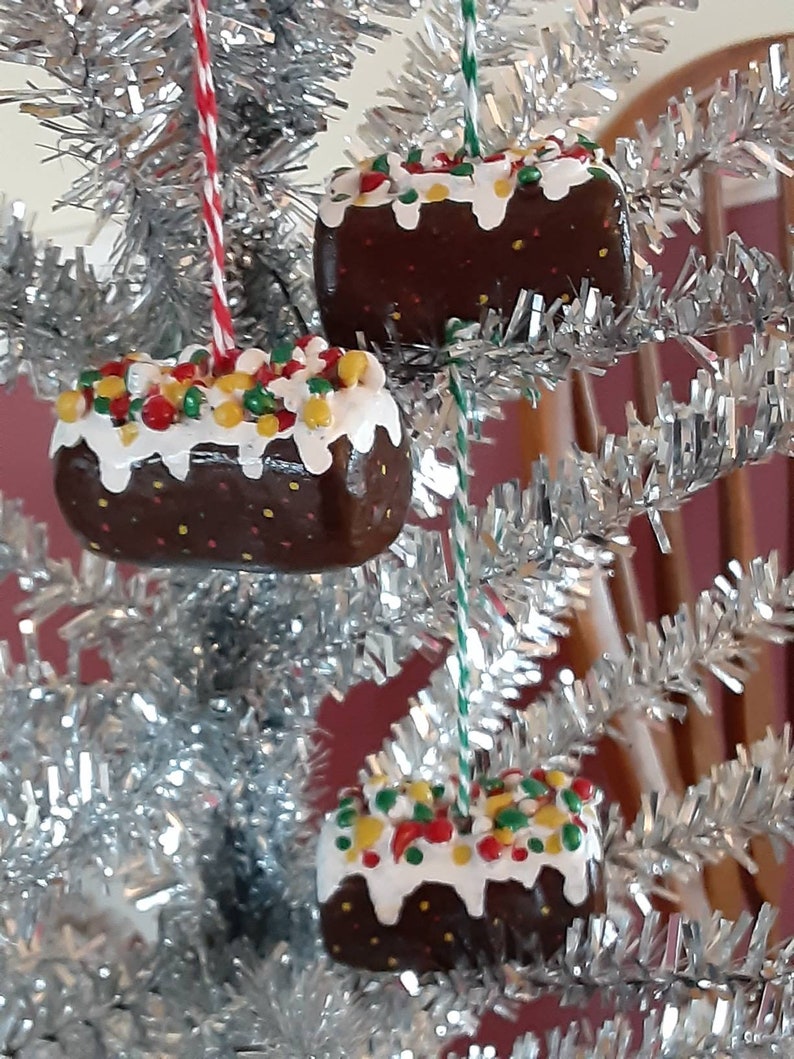 Peut inclure: Trois d&eacute;corations de No&euml;l en forme de g&acirc;teaux au chocolat avec un gla&ccedil;age blanc et des vermicelles color&eacute;s. Les d&eacute;corations sont suspendues &agrave; un sapin de No&euml;l en tinsel argent&eacute;.