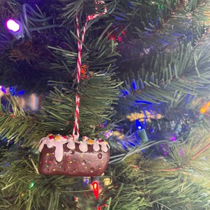 Peut inclure: Une d&eacute;coration de No&euml;l en forme de g&acirc;teau avec des p&eacute;pites color&eacute;es, de couleur marron et blanche. La d&eacute;coration est suspendue &agrave; une ficelle rouge et blanche sur un sapin de No&euml;l vert.