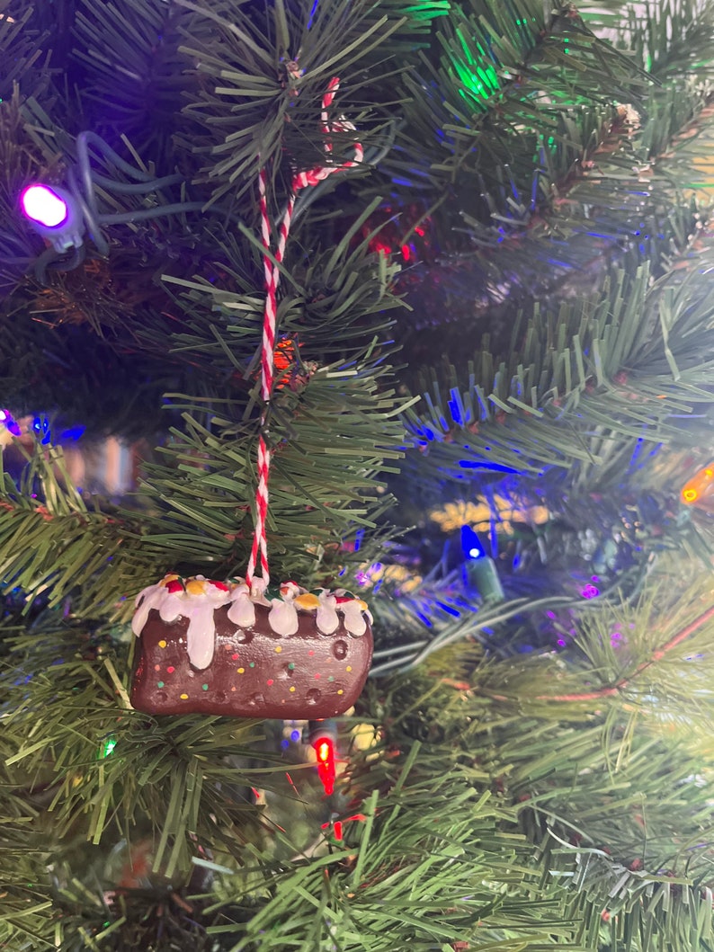 Peut inclure: Une d&eacute;coration de No&euml;l en forme de g&acirc;teau avec des p&eacute;pites color&eacute;es, de couleur marron et blanche. La d&eacute;coration est suspendue &agrave; une ficelle rouge et blanche sur un sapin de No&euml;l vert.