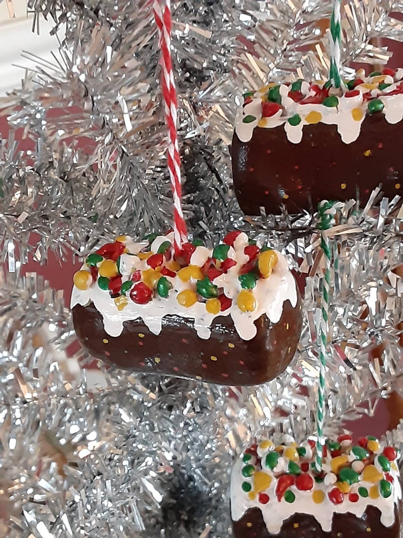 Peut inclure: Trois d&eacute;corations de No&euml;l en forme de g&acirc;teau au chocolat avec gla&ccedil;age blanc et p&eacute;pites color&eacute;es. Les d&eacute;corations sont suspendues &agrave; un sapin de No&euml;l en tinsel argent&eacute;.