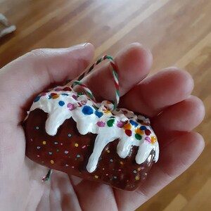 Peut inclure: Une petite d&eacute;coration de g&acirc;teau peinte &agrave; la main, de couleur marron et blanche, avec des vermicelles color&eacute;s. La d&eacute;coration est munie d'une ficelle pour l'accrocher.