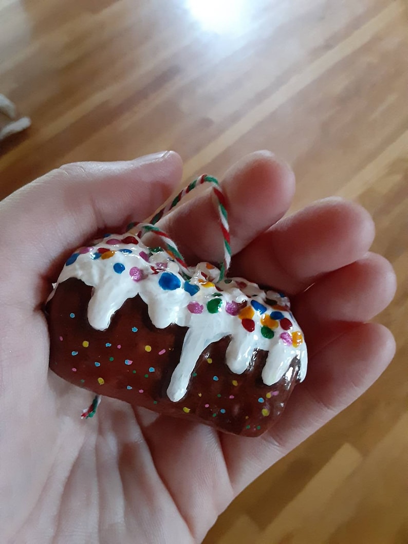 Peut inclure: Une petite d&eacute;coration de g&acirc;teau peinte &agrave; la main, de couleur marron et blanche, avec des vermicelles color&eacute;s. La d&eacute;coration est munie d'une ficelle pour l'accrocher.