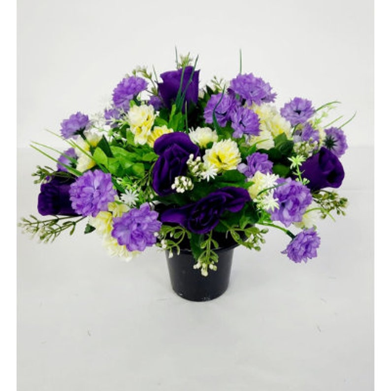 May include: A purple and white artificial flower arrangement in a black pot. The arrangement features roses, carnations, and other small flowers.