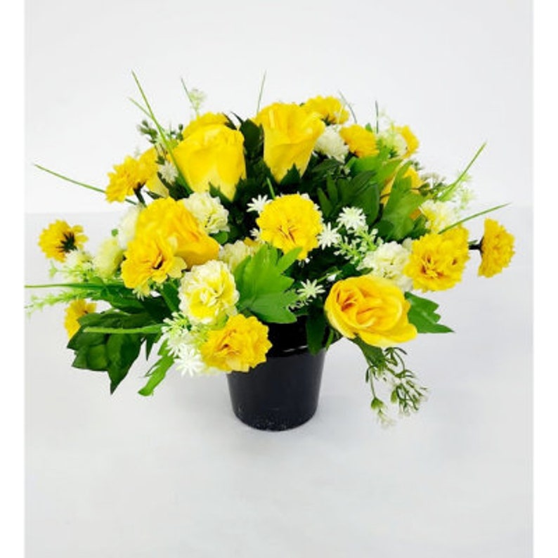 May include: A yellow artificial flower arrangement in a black pot. The arrangement features roses, carnations, and other small white flowers.