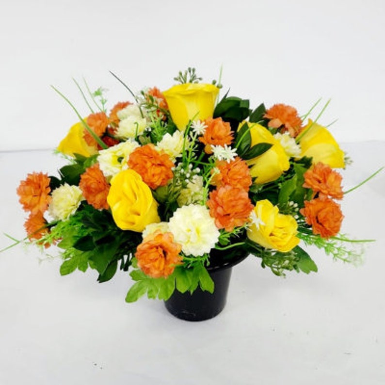 May include: A yellow and orange artificial flower arrangement in a black pot. The arrangement features roses, carnations, and other small flowers.