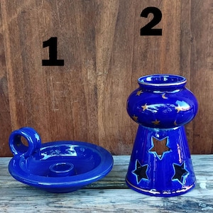 Può includere: Due portacandele in ceramica blu. Uno è una piccola ciotola con manico. L'altro è un portacandele alto e cilindrico con ritagli a forma di stella e una parte superiore arrotondata decorata con stelle e lune dorate.